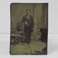 Antique Tintype Photo: Man, standing, leaning one arm on the back of leather back chair with padded arms and wood frame: Front View - Click to enlarge